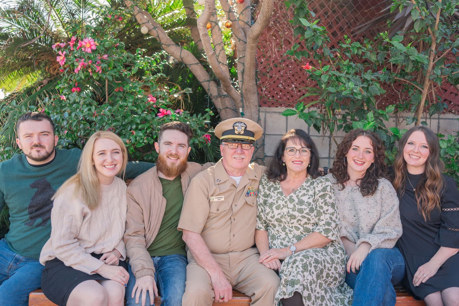 A group sits together on a wooden bench in a garden setting with pink flowers and greenery in the background.
