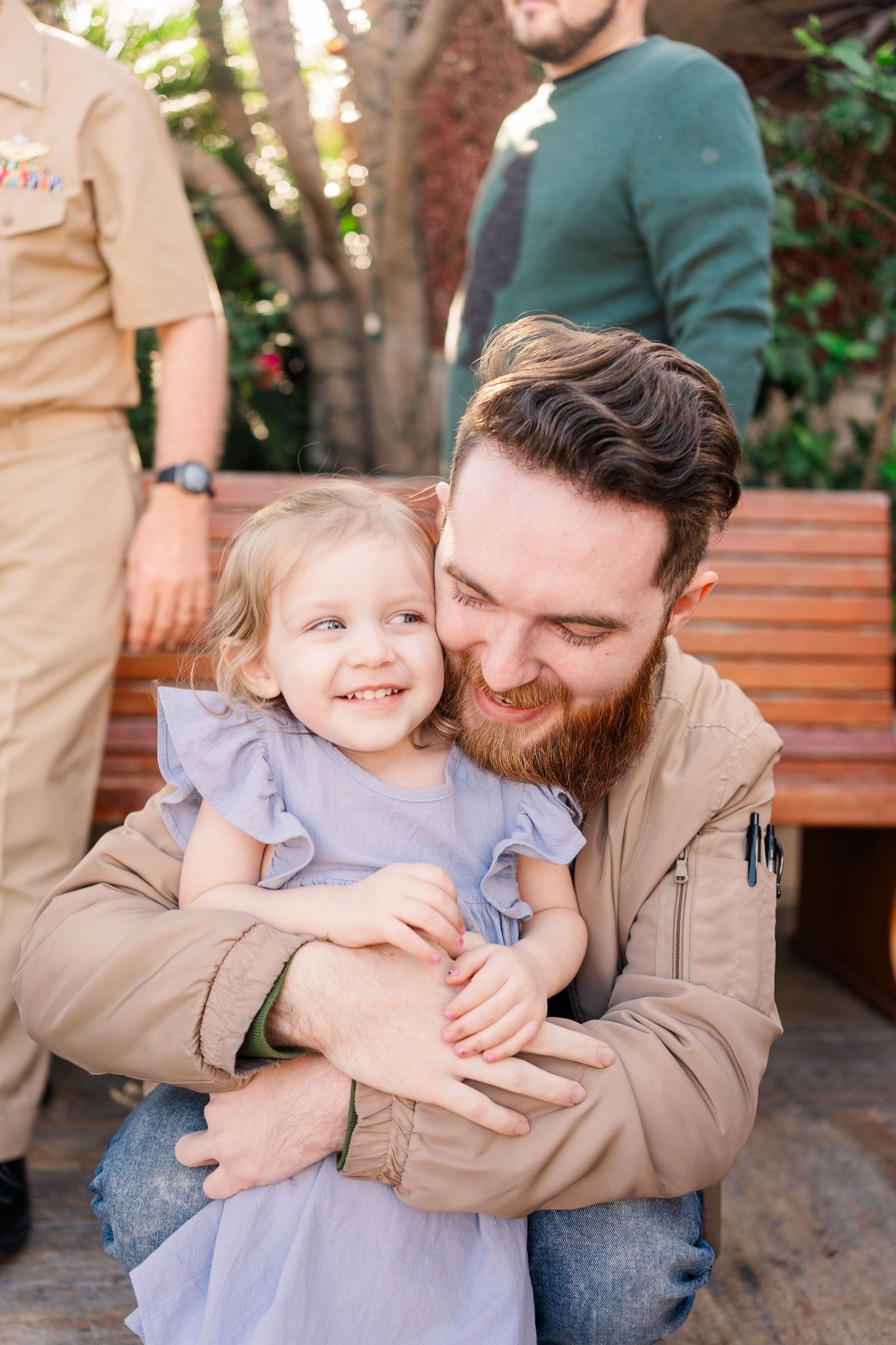 A bearded parent in a tan jacket shares a joyful moment with a small child in a blue dress outdoors.