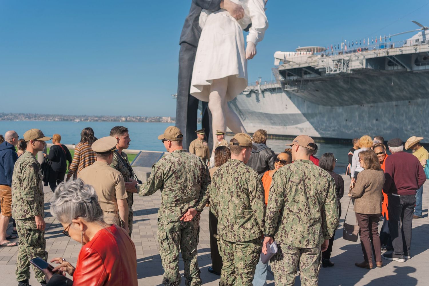 Military service members in camouflage uniforms gather on a pier next to a naval ship under bright blue skies.