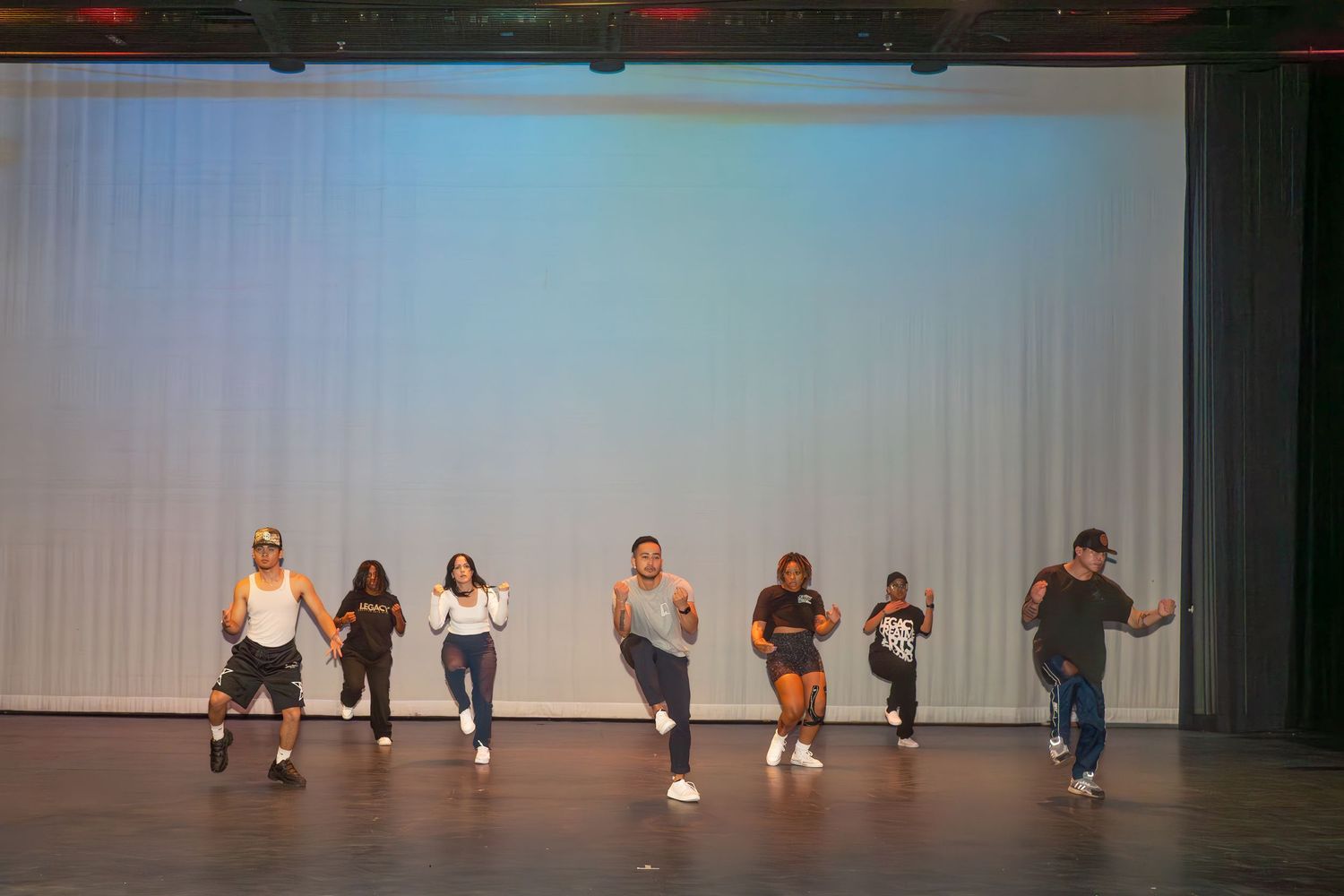 A group of dancers perform in sync on a stage with white backdrop during a rehearsal or performance.