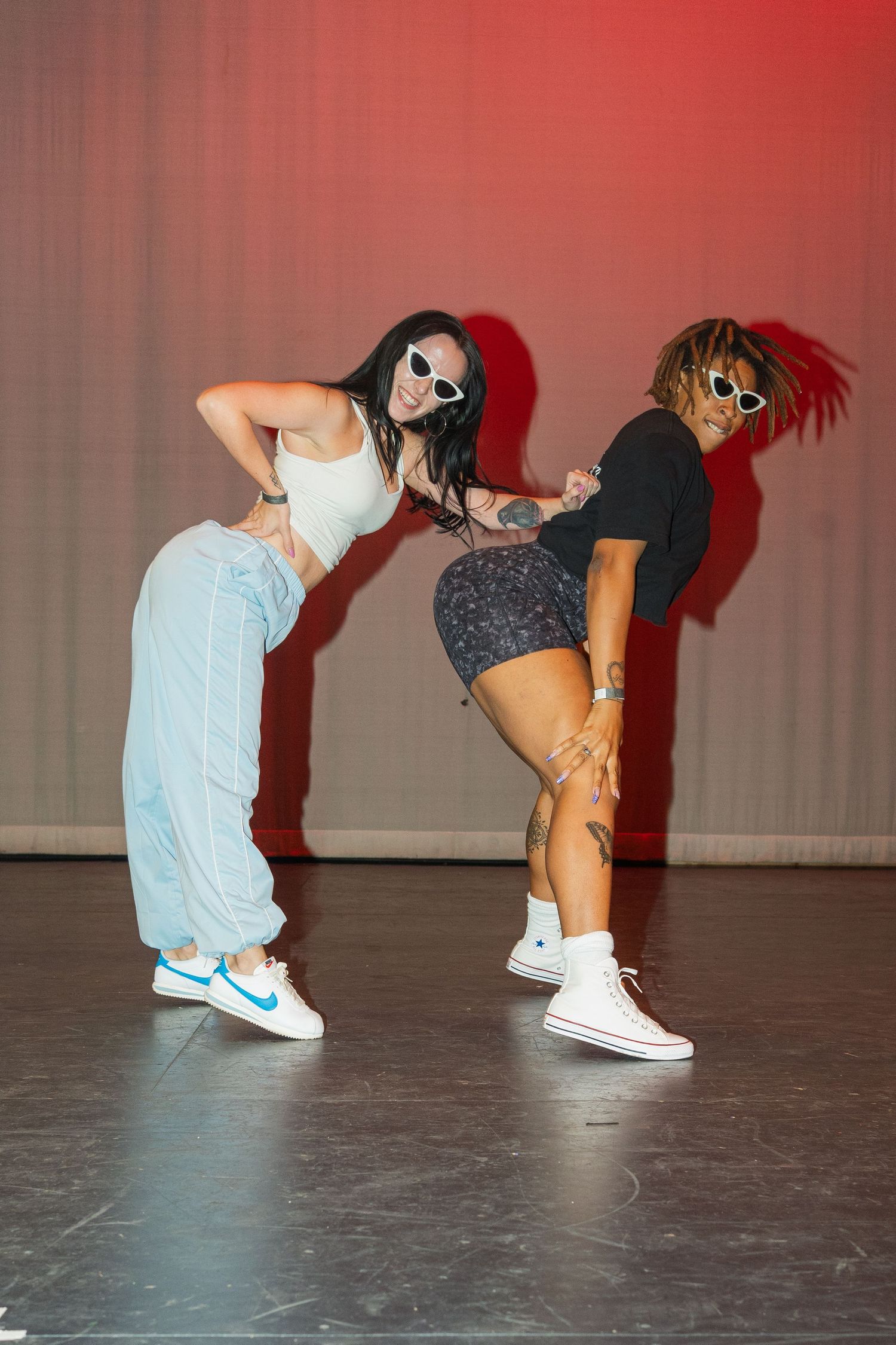 Two dancers perform energetic hip hop moves wearing casual street style clothing on a dark stage with red lighting.