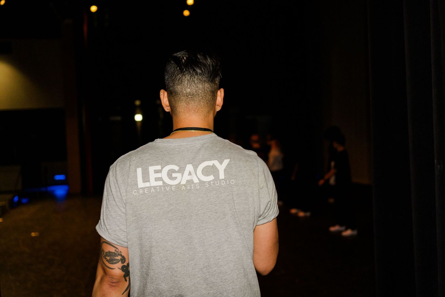 Person wearing a gray t-shirt with LEGACY text on the back stands in a dimly lit environment at night.