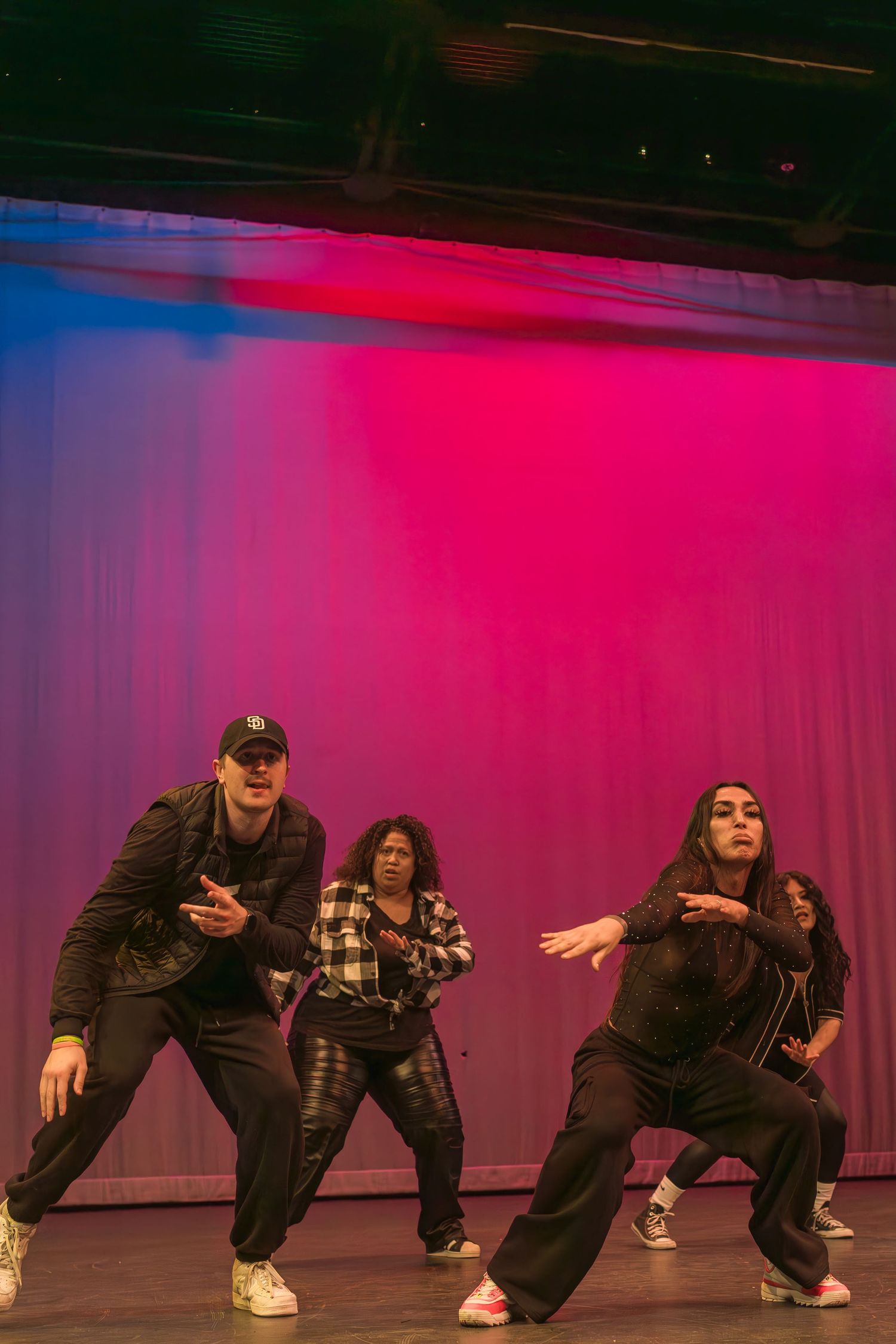 Three dancers perform energetic hip hop choreography on stage with vibrant pink and blue lighting effects.