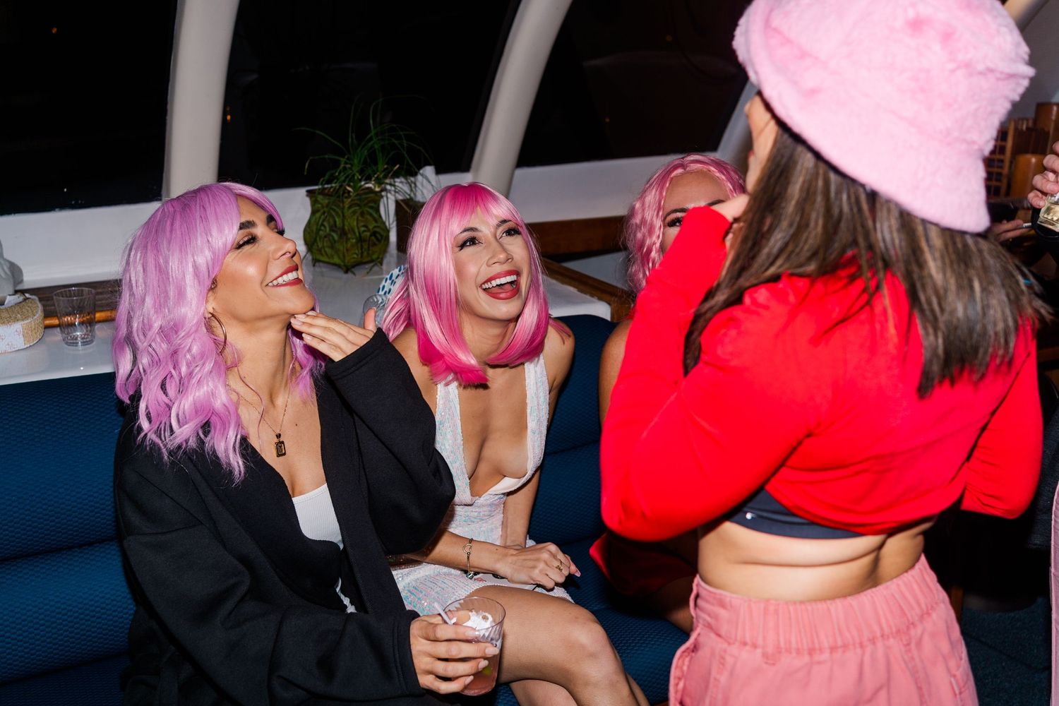 Candid nightlife scene showing people with colorful pink hair laughing together on blue seating at an event.
