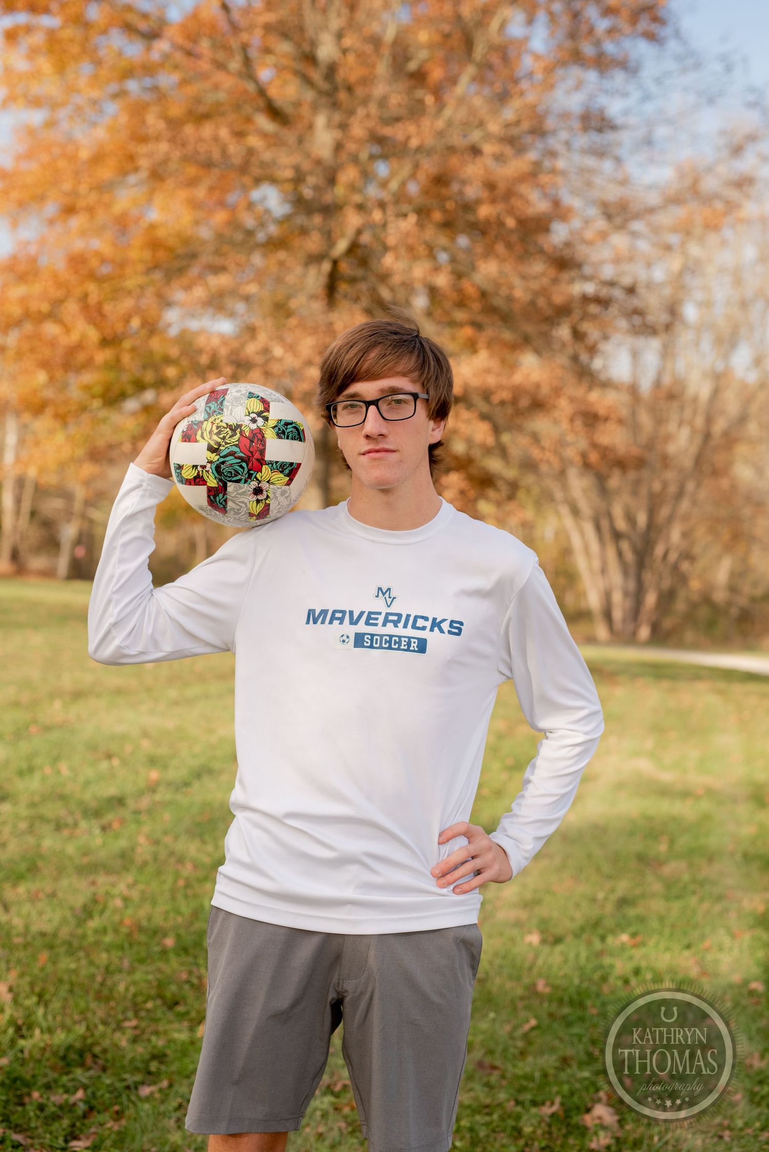 Person holds soccer ball while posing in Mavericks shirt against fall scenery.