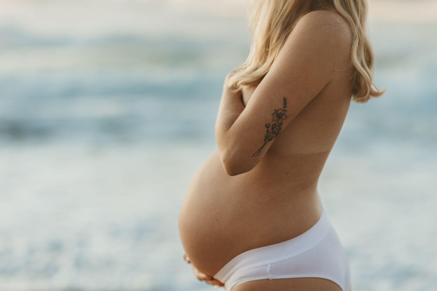 Pregnant woman, blonde hair, holding her stomach, staring out at the ocean
