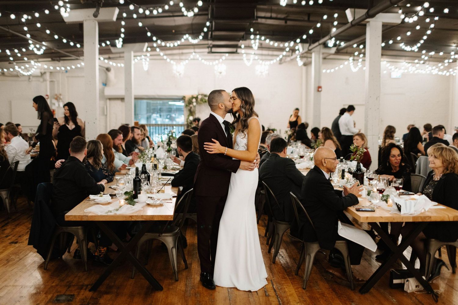 Wedding reception in an industrial venue with string lights and guests seated at long tables.