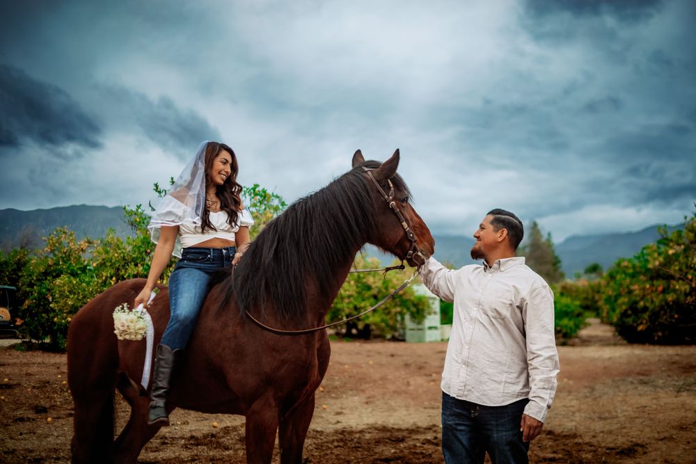 Engaged couple and a majestic dark brown horse stand together against a dramatic cloudy mountain landscape in Riverside California