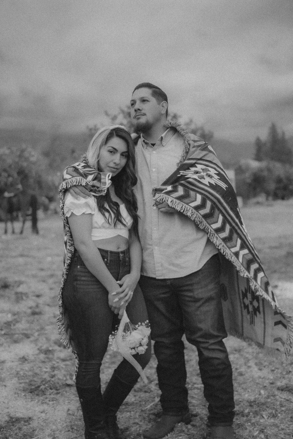 A romantic black and white portrait of a couple embracing outdoors, wrapped in a patterned blanket in a rustic setting.
