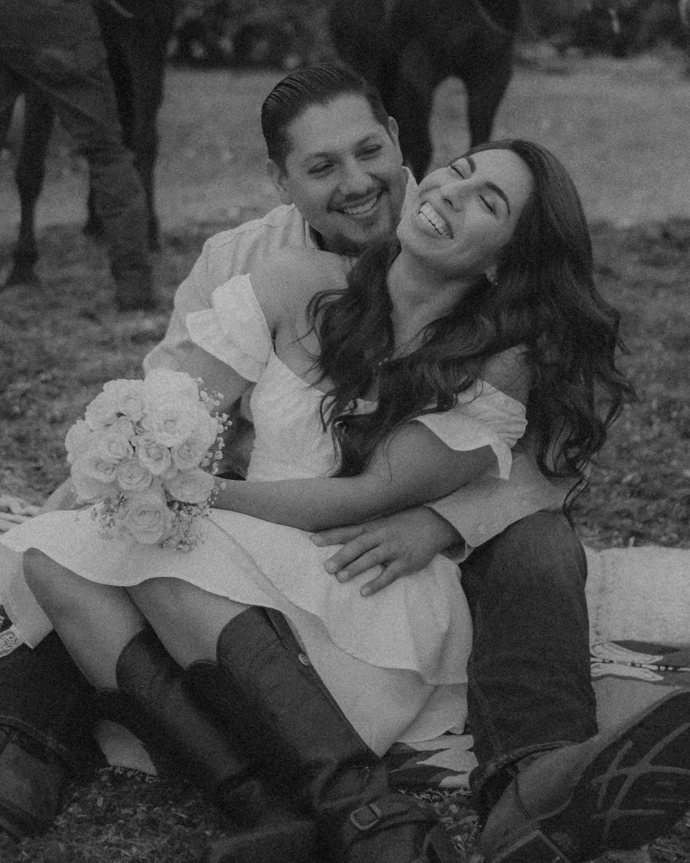 A black and white photo of a couple in casual attire sharing a happy moment outdoors on a blanket with flowers nearby.