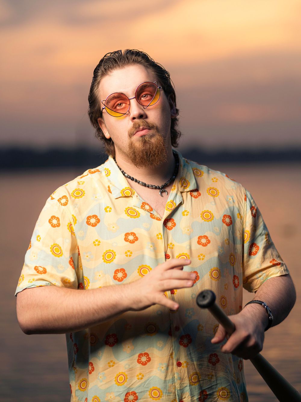 Person in floral print shirt poses with walking stick during golden hour by waterfront.