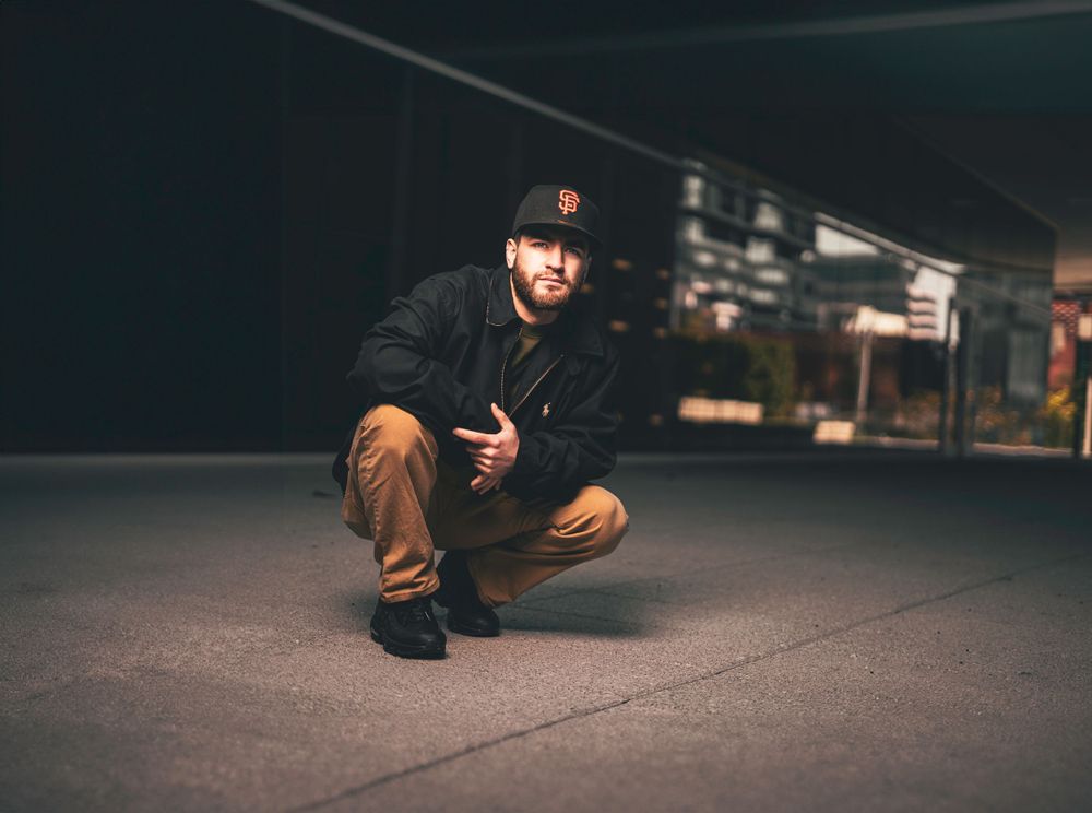 Urban portrait of a person in casual wear crouching on the ground in a dark indoor setting.