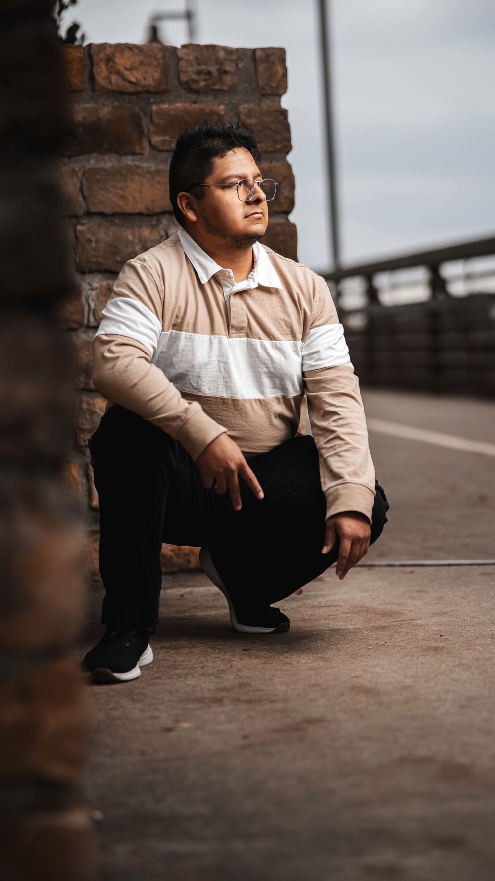 A person in a beige and white striped sweater crouches against a brick wall in a contemplative urban portrait.