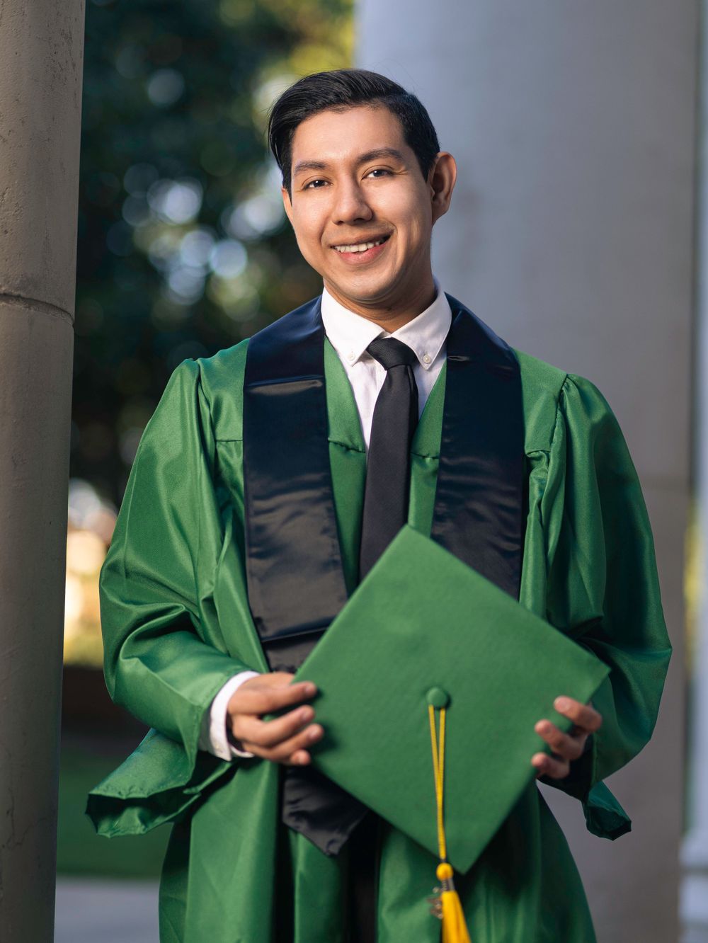 Graduate in green and black academic regalia holds diploma cover while standing by pillar outdoors.