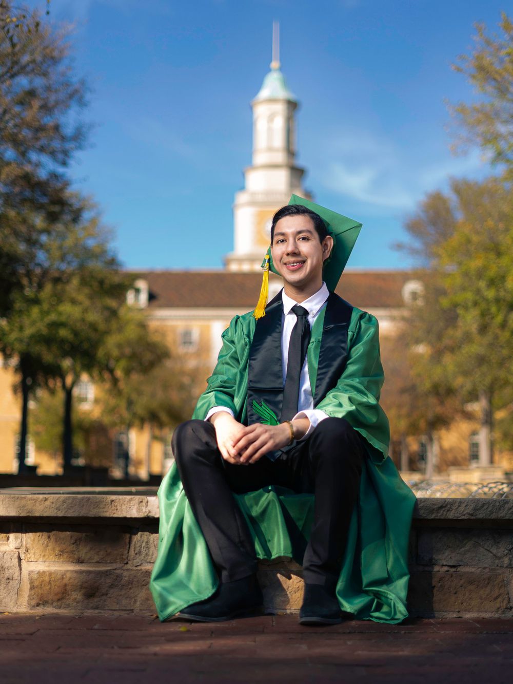 Graduate in green cap and gown sitting in front of a university building with white steeple.