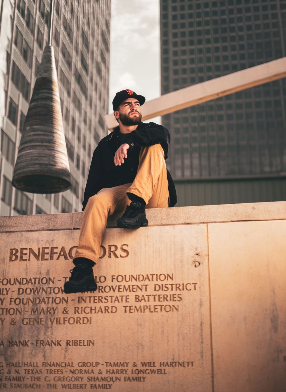 Person sitting on a stone wall marked 'Benefactors' with modern skyscrapers in the background.