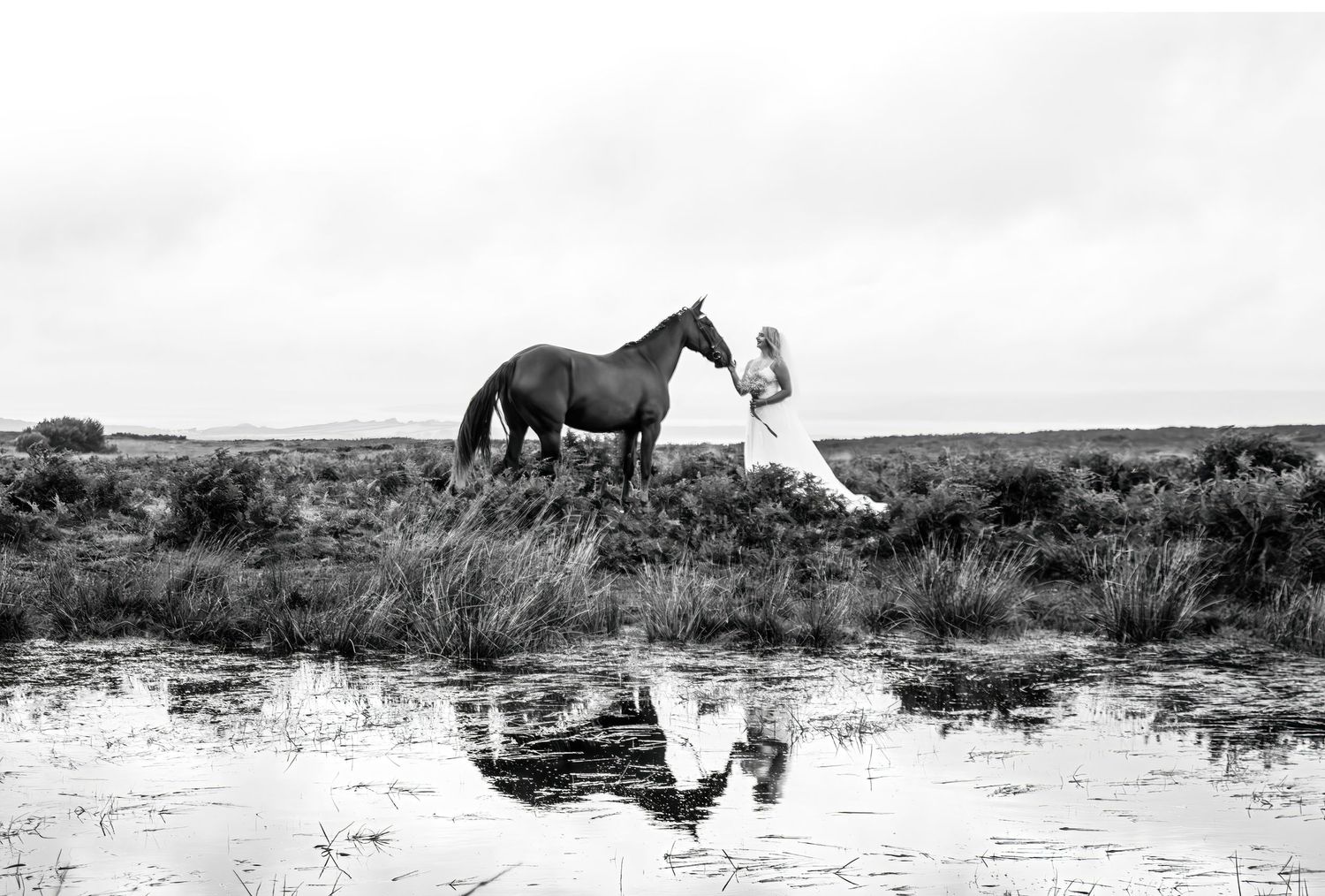 A white dress stands out against dark horse and reflection in water on misty moors.