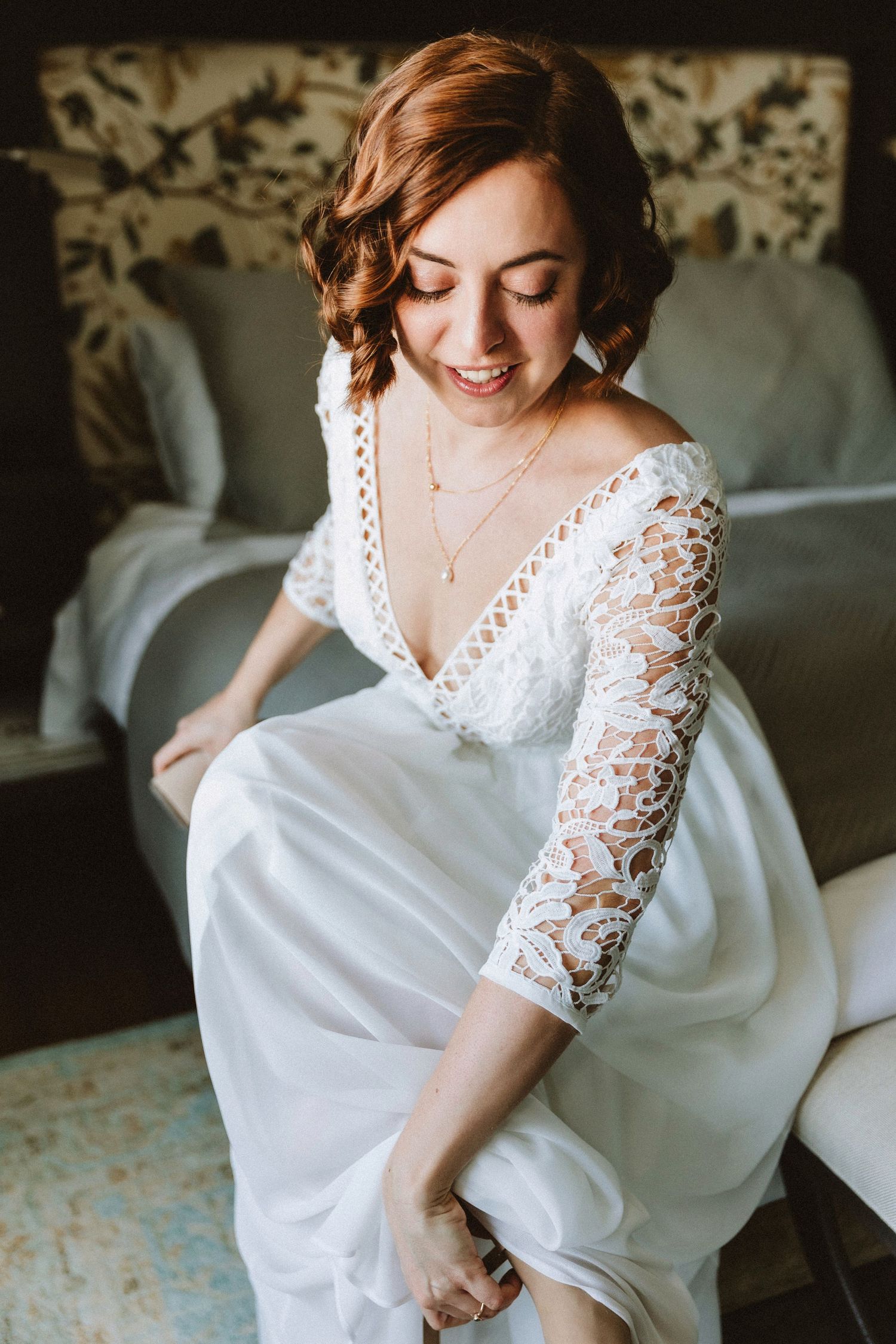 Natural light illuminates a bride in lace sleeve wedding dress sitting on patterned chair.