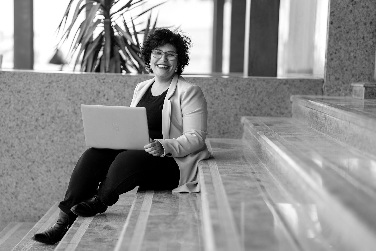 Person sitting on steps with laptop wearing casual attire in black and white photography.