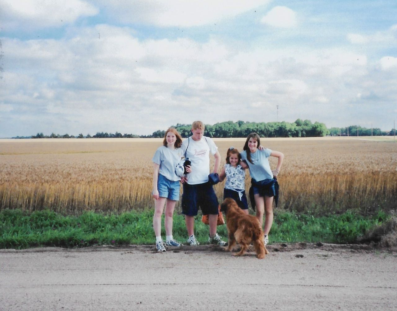 A family group with their dog posing on a dirt road alongside a wheat field on a cloudy day.