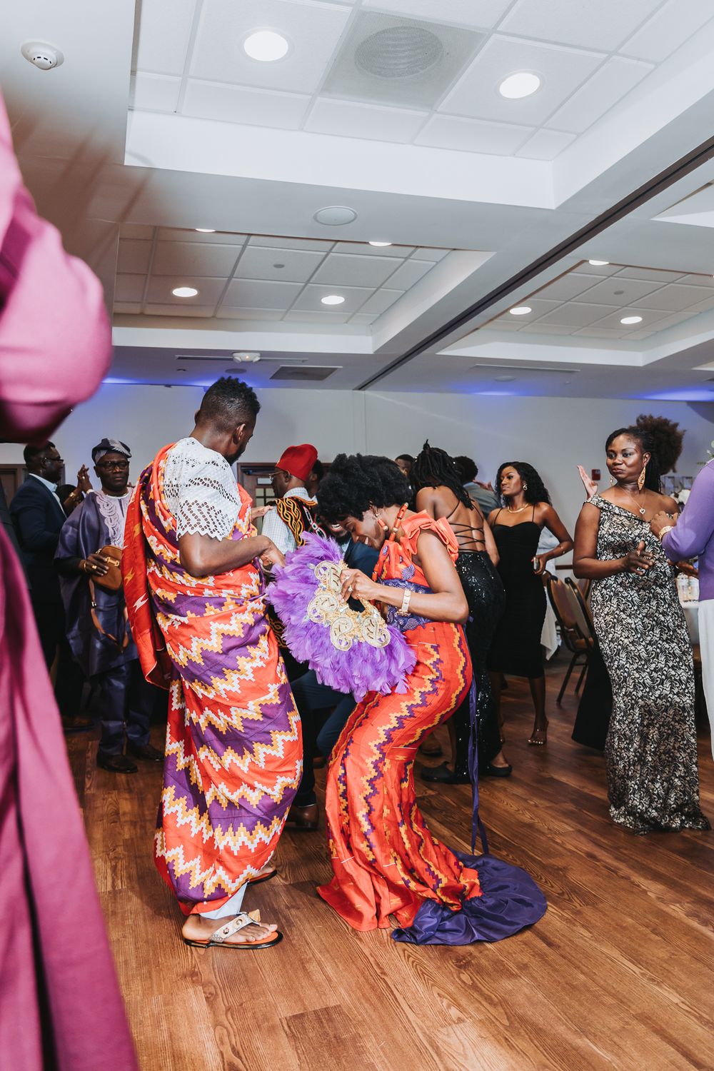 People dancing at a celebration wearing vibrant patterned dresses in orange and red on a wooden dance floor.