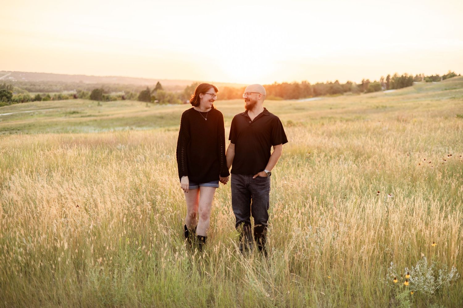 A couple stands together in a golden meadow at sunset with rolling hills in the background.