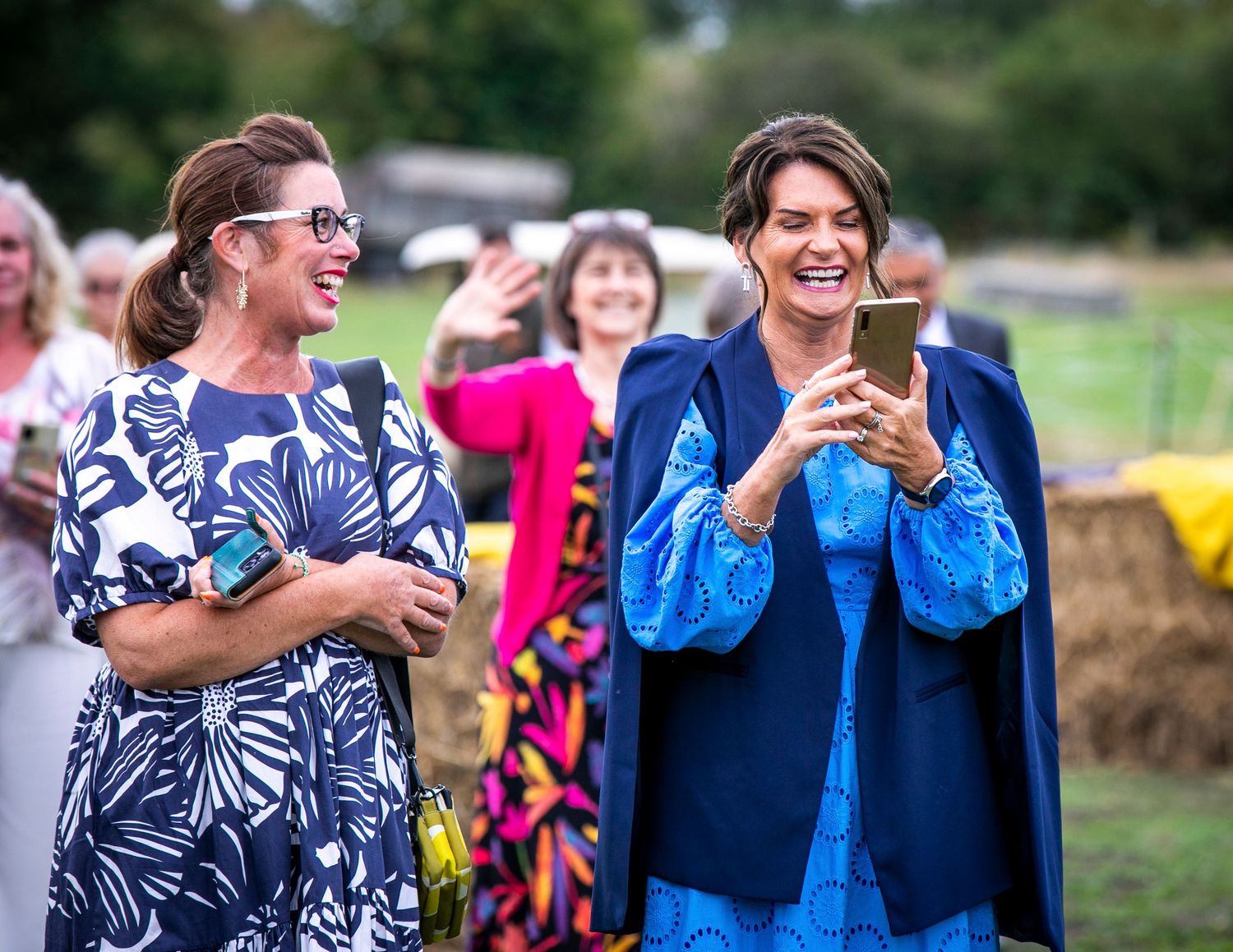 Two wedding guests sharing a laugh while looking at their phones during an outdoor celebration.