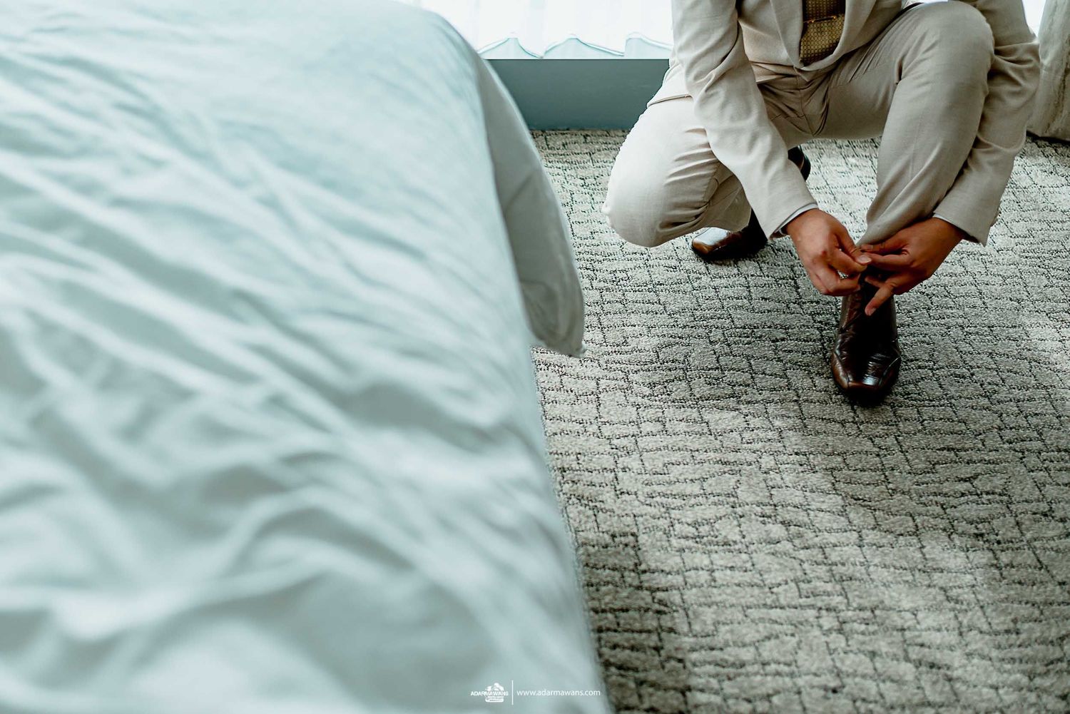 A person in a light colored suit sits on carpet while putting on formal shoes.