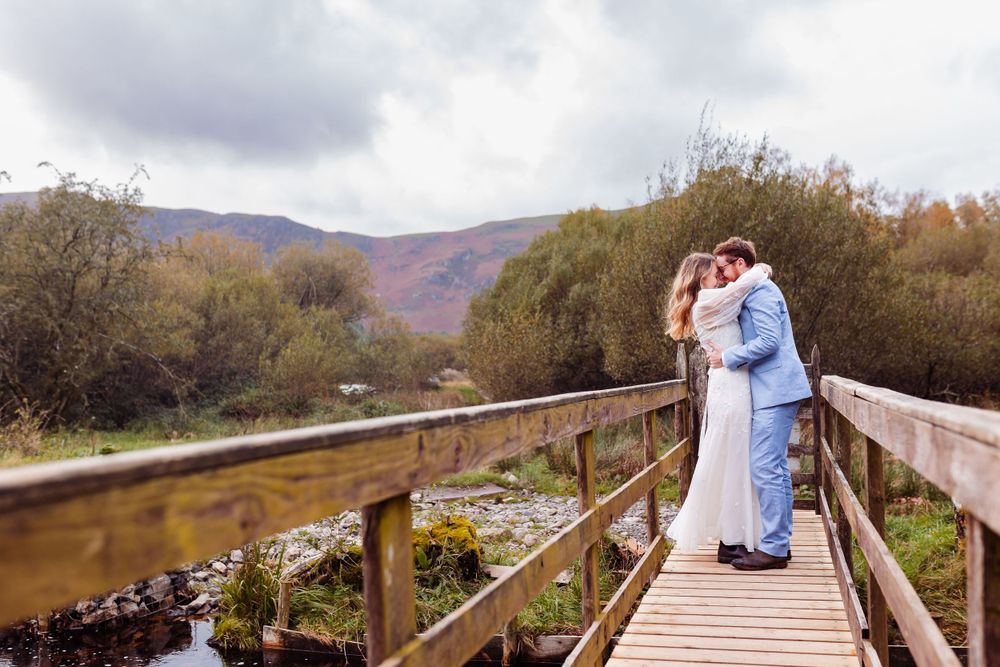 Couple in light blue outfits embracing on wooden bridge with autumn mountains.