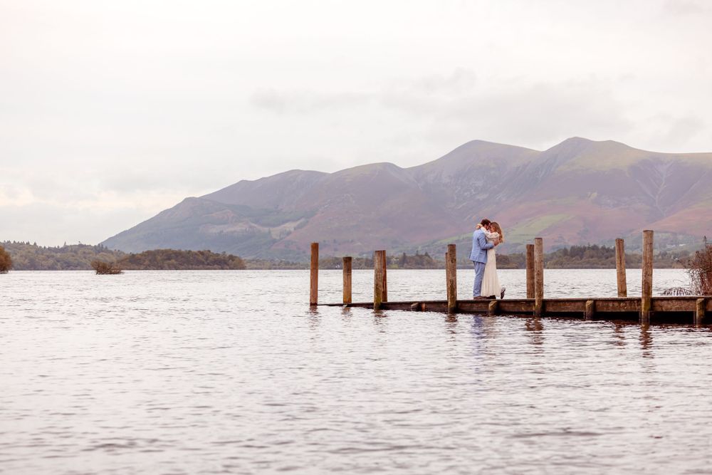 A couple shares an intimate moment on a wooden jetty with mountains in the distance.