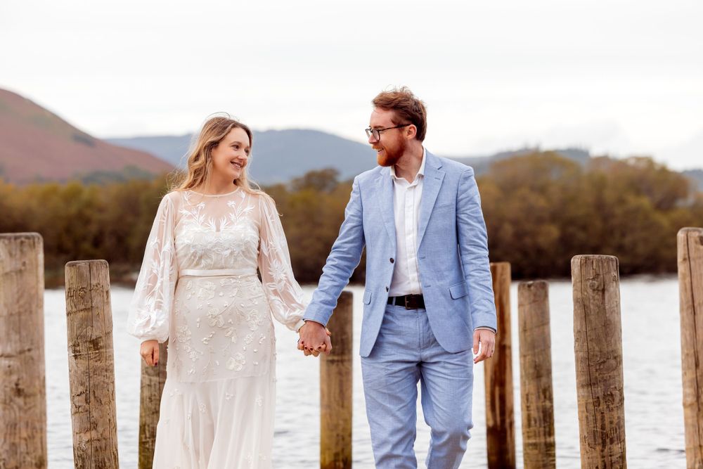 A couple in wedding attire holds hands while walking on a wooden pier with mountains behind them.
