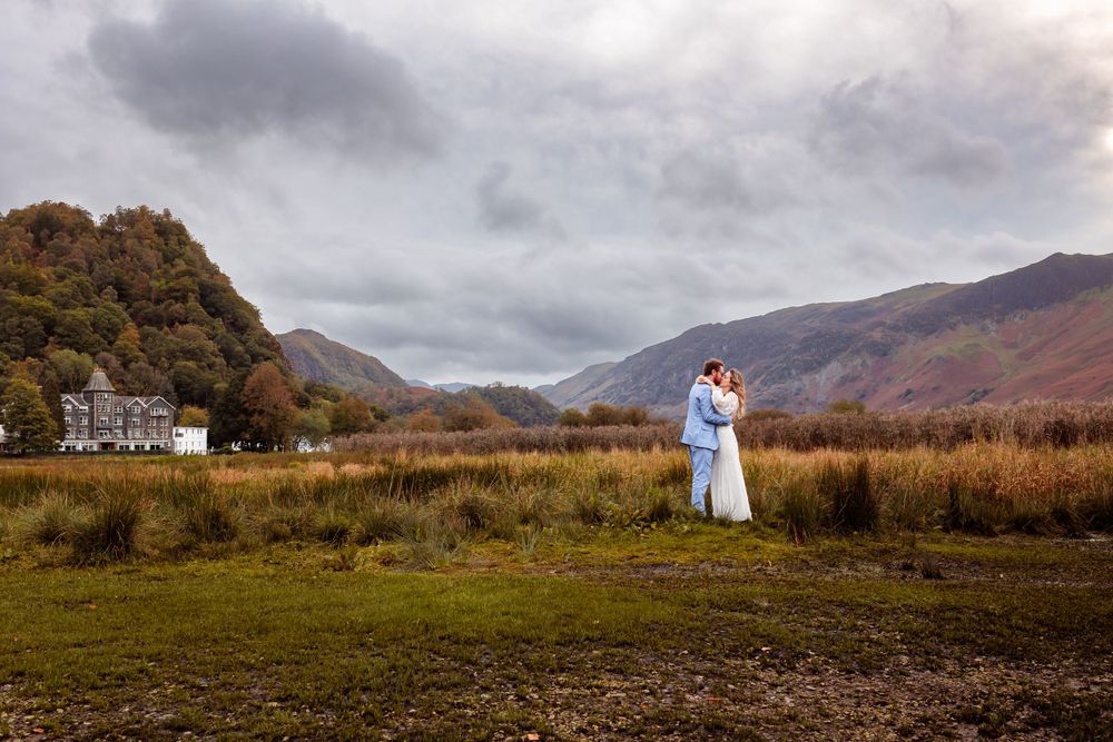 A couple embraces on a grassy hill with mountains and moody skies in the Scottish Highlands landscape.