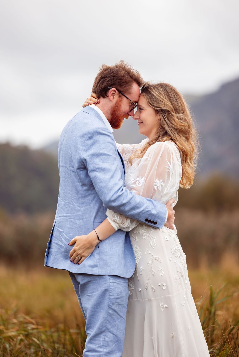 Couple in blue suit and white dress sharing intimate moment against mountain backdrop.