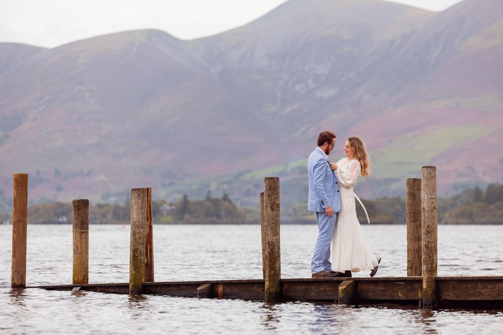 A couple stands on a wooden pier overlooking a lake surrounded by misty mountains.