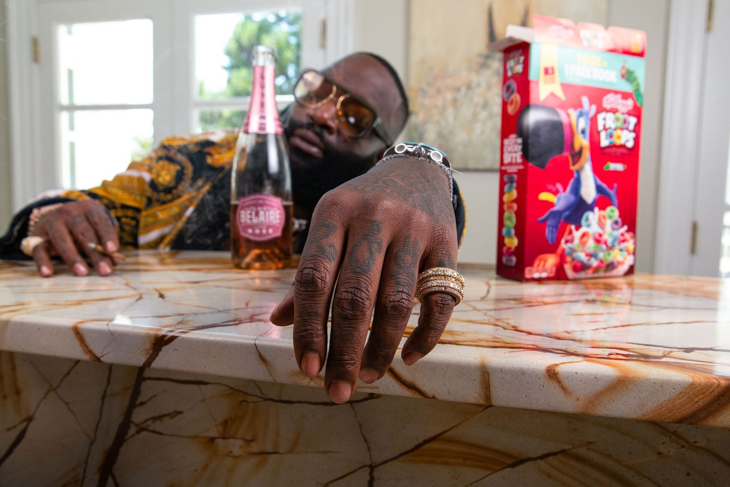 Rick Ross sitting at a breakfast bar posed with a Belaire champagne bottle and cereal box.