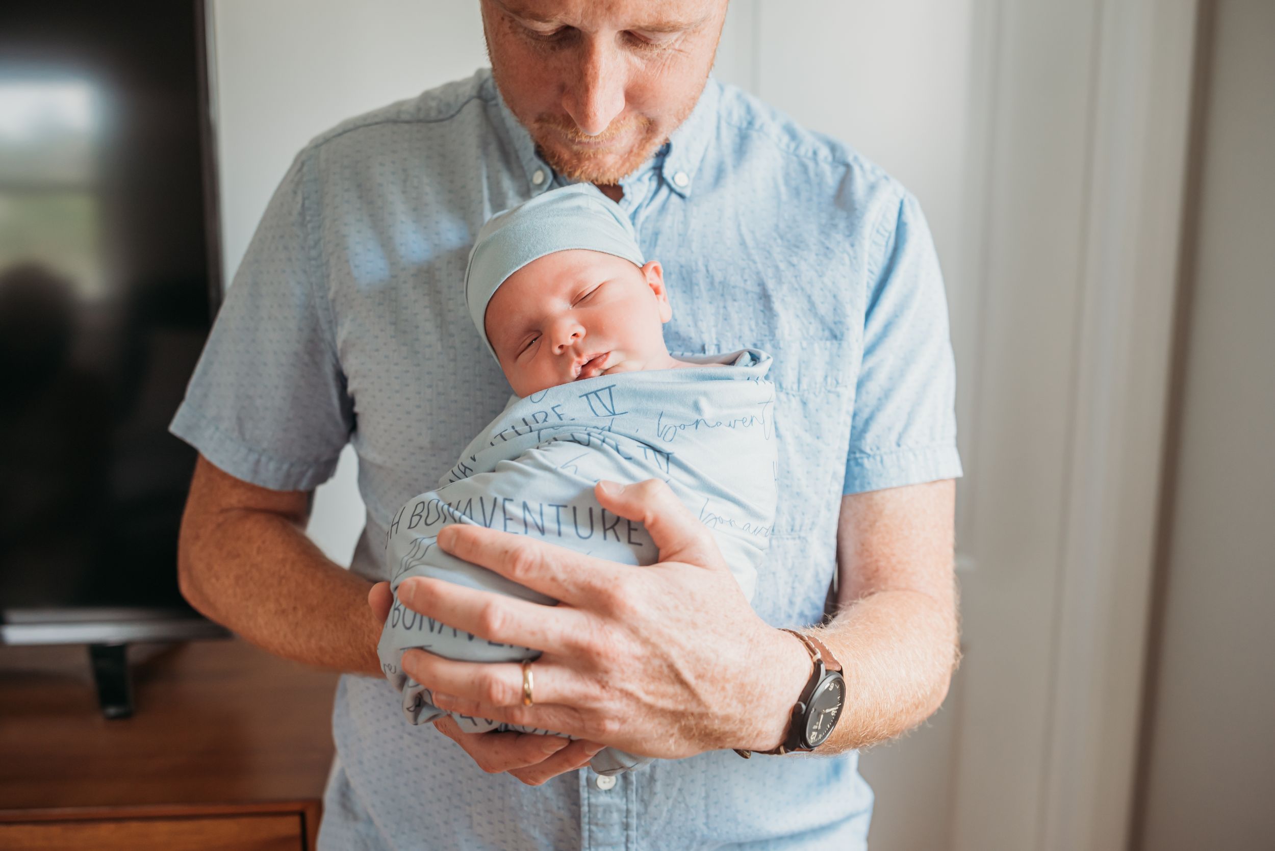 Dad holds his new baby boy wrapped in his custom name blue swaddle in front of his body and smiles down at him.