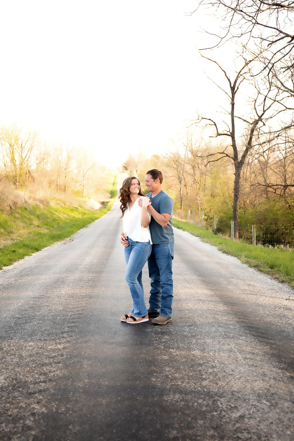 A couple embraces on a rural road during sunset with trees and golden light in the background.