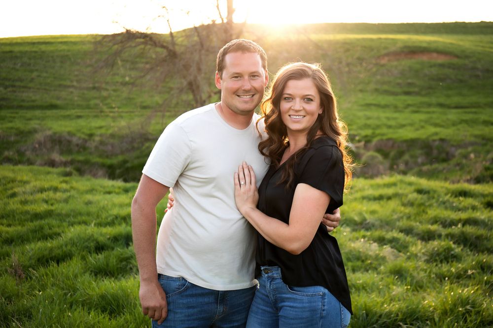 A couple poses together smiling in a lush green field during golden hour.