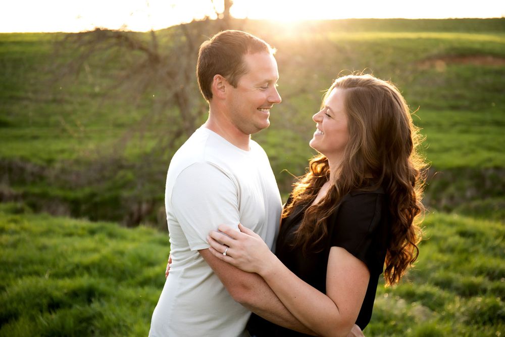 A happy couple embraces while sharing a laugh in a sunlit grassy field.
