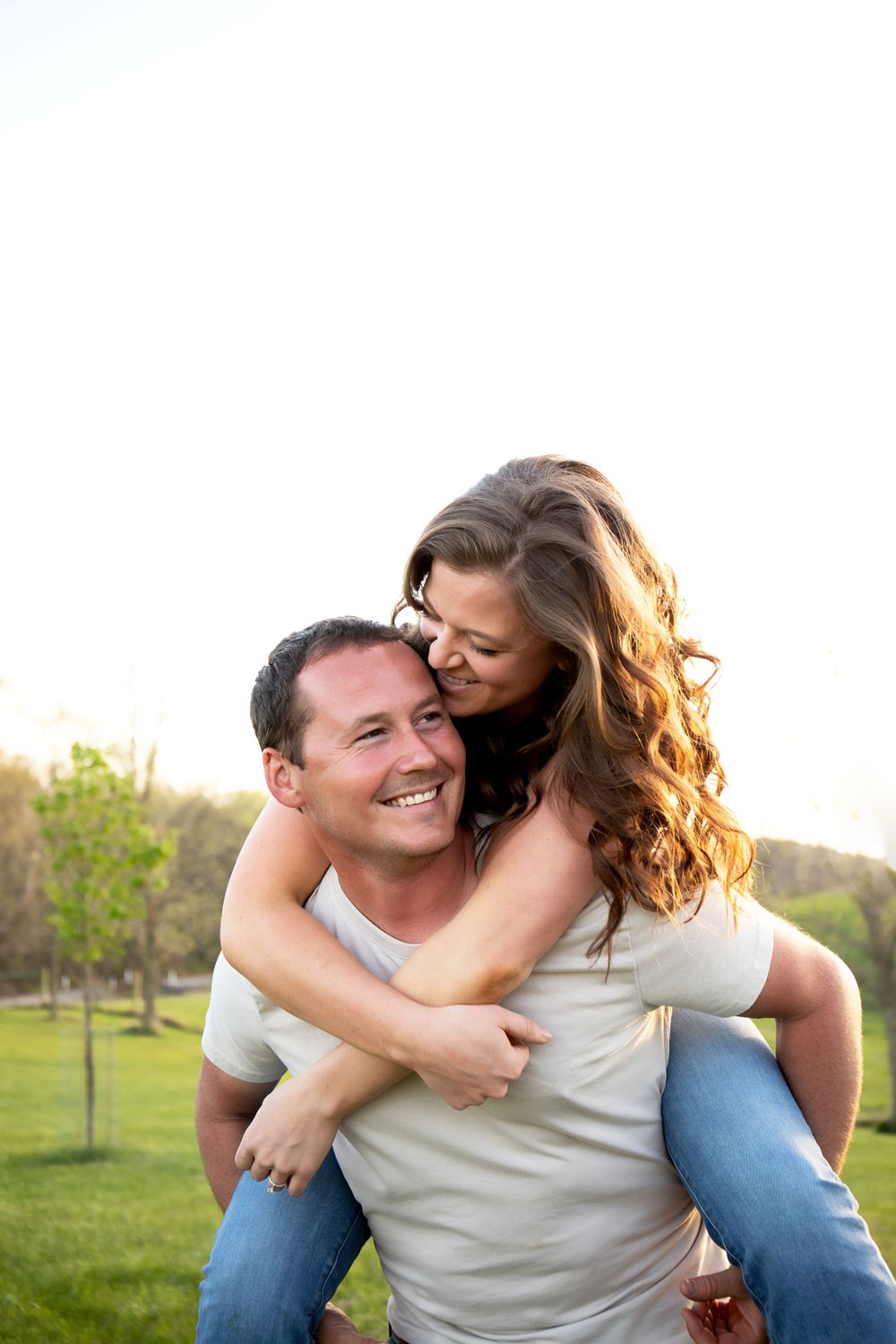 A couple shares a fun piggyback ride moment outdoors in a grassy park.
