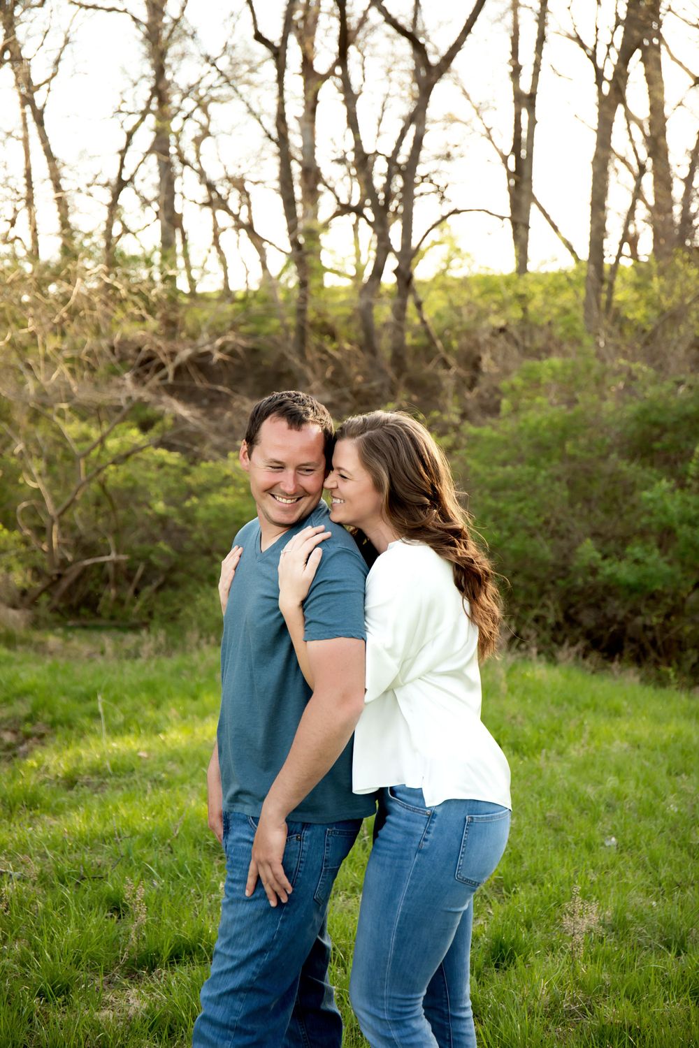 A playful couple shares a laugh while embracing in a wooded area during sunset.