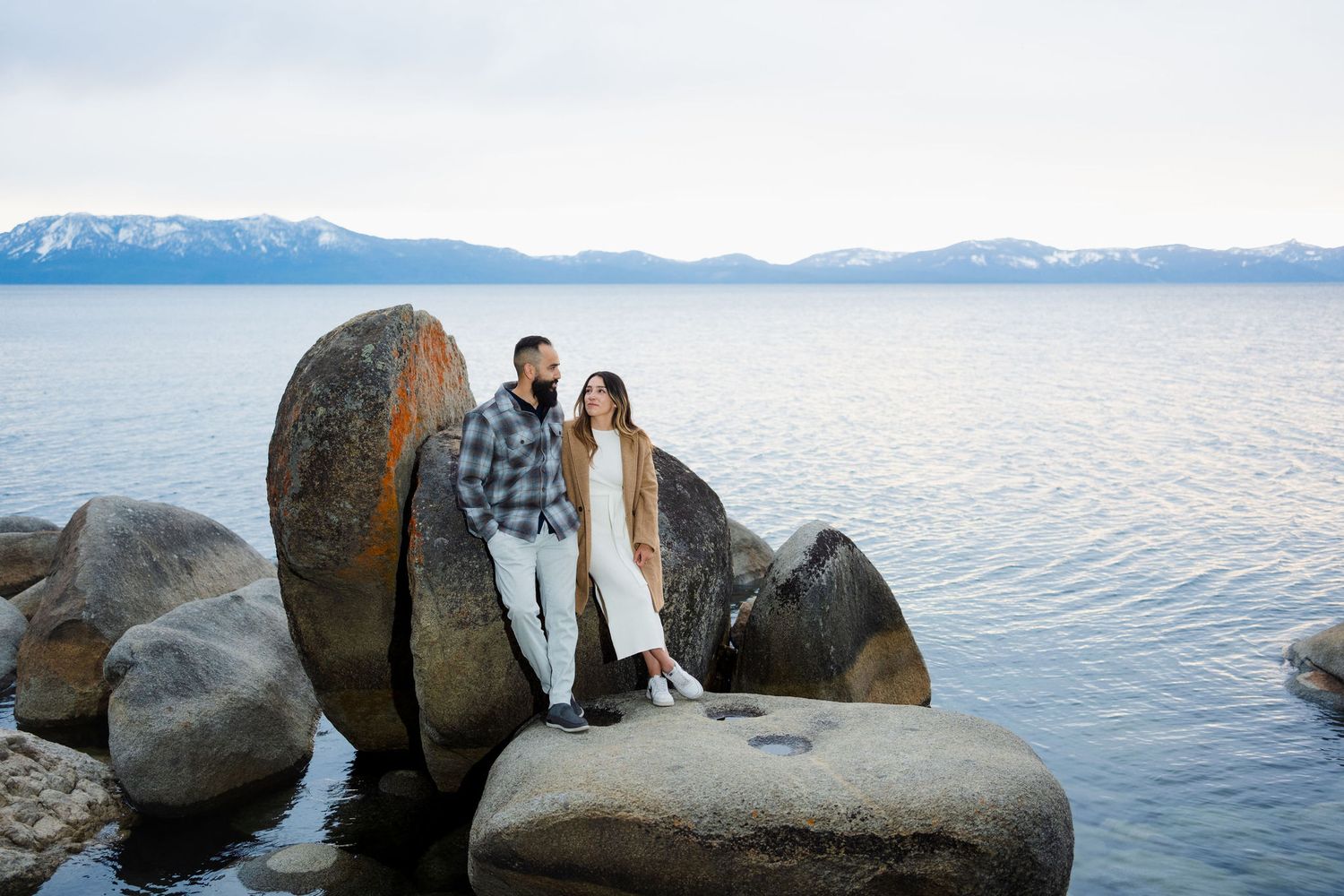 A couple stands on large boulders at Lake Tahoe with snow-capped mountains visible in the background.