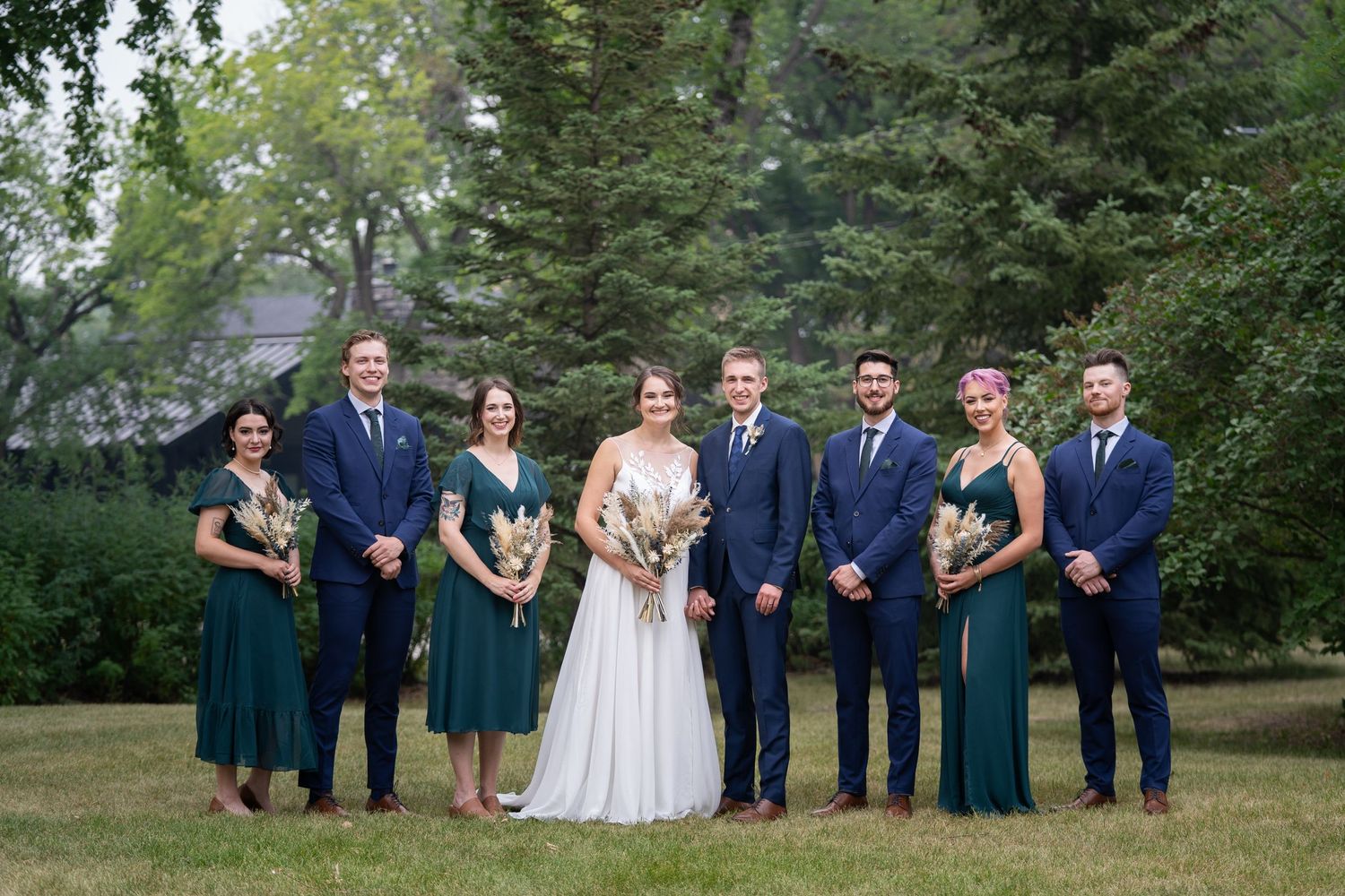 Wedding party in navy suits and teal dresses pose formally on grass with evergreen backdrop.