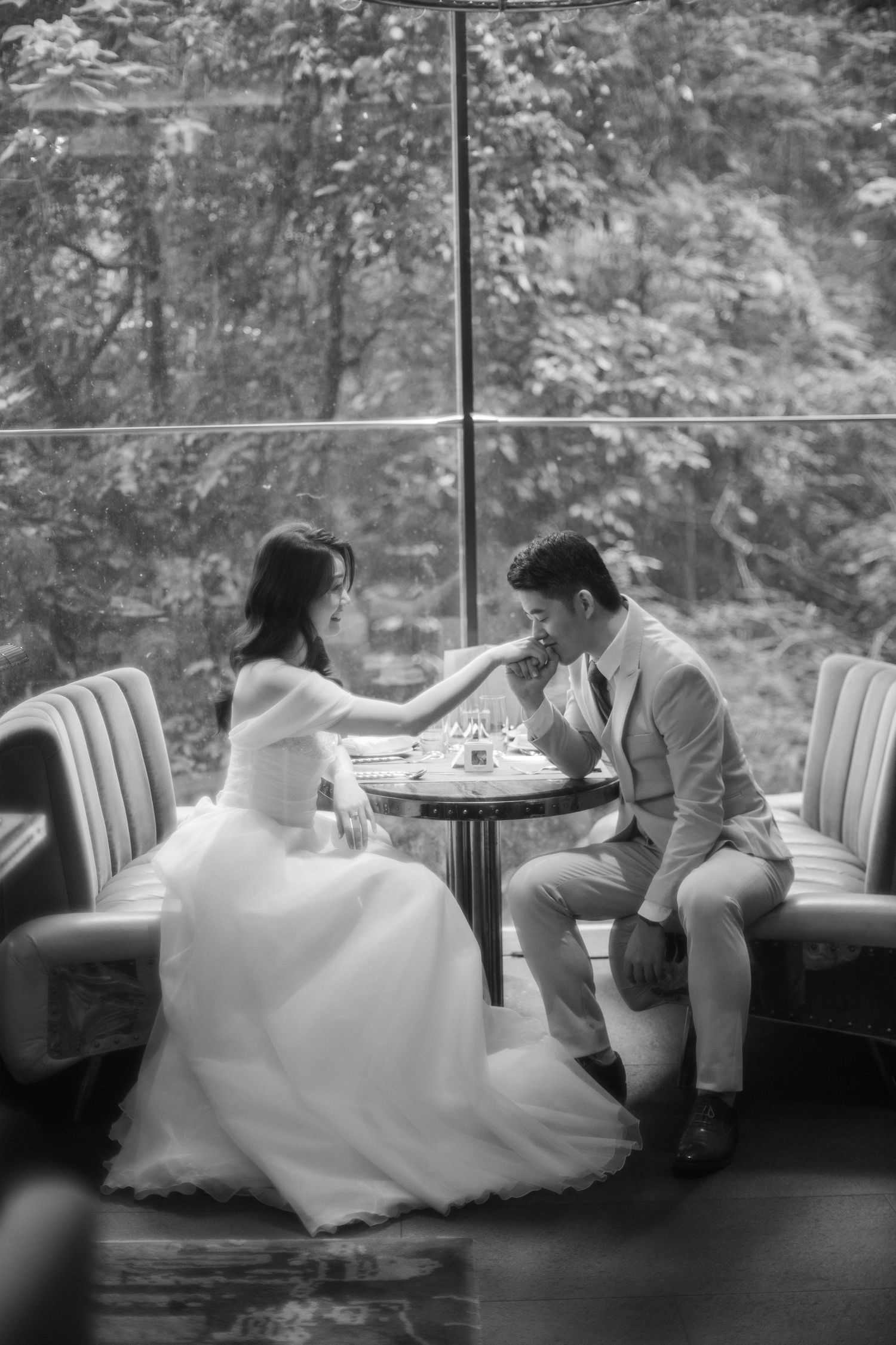 A tender black and white wedding photo of a couple sharing an intimate moment at a table by large windows.