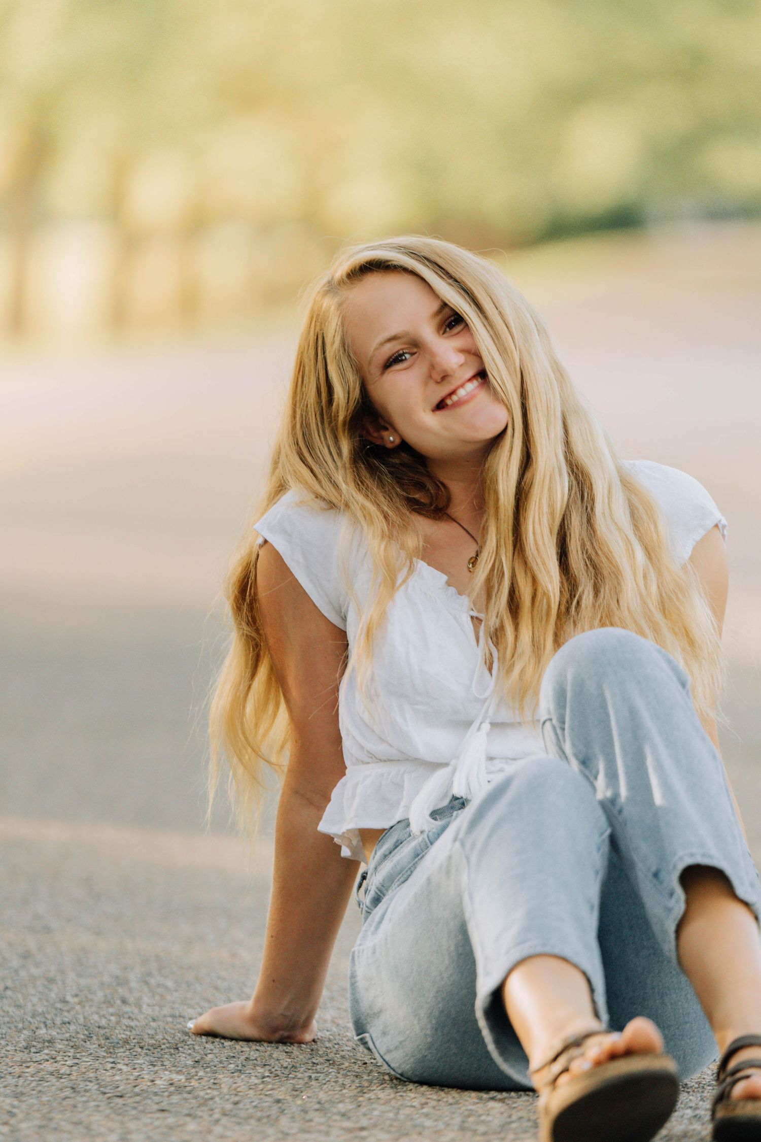 Person in white top and light jeans sitting casually on ground outdoors.