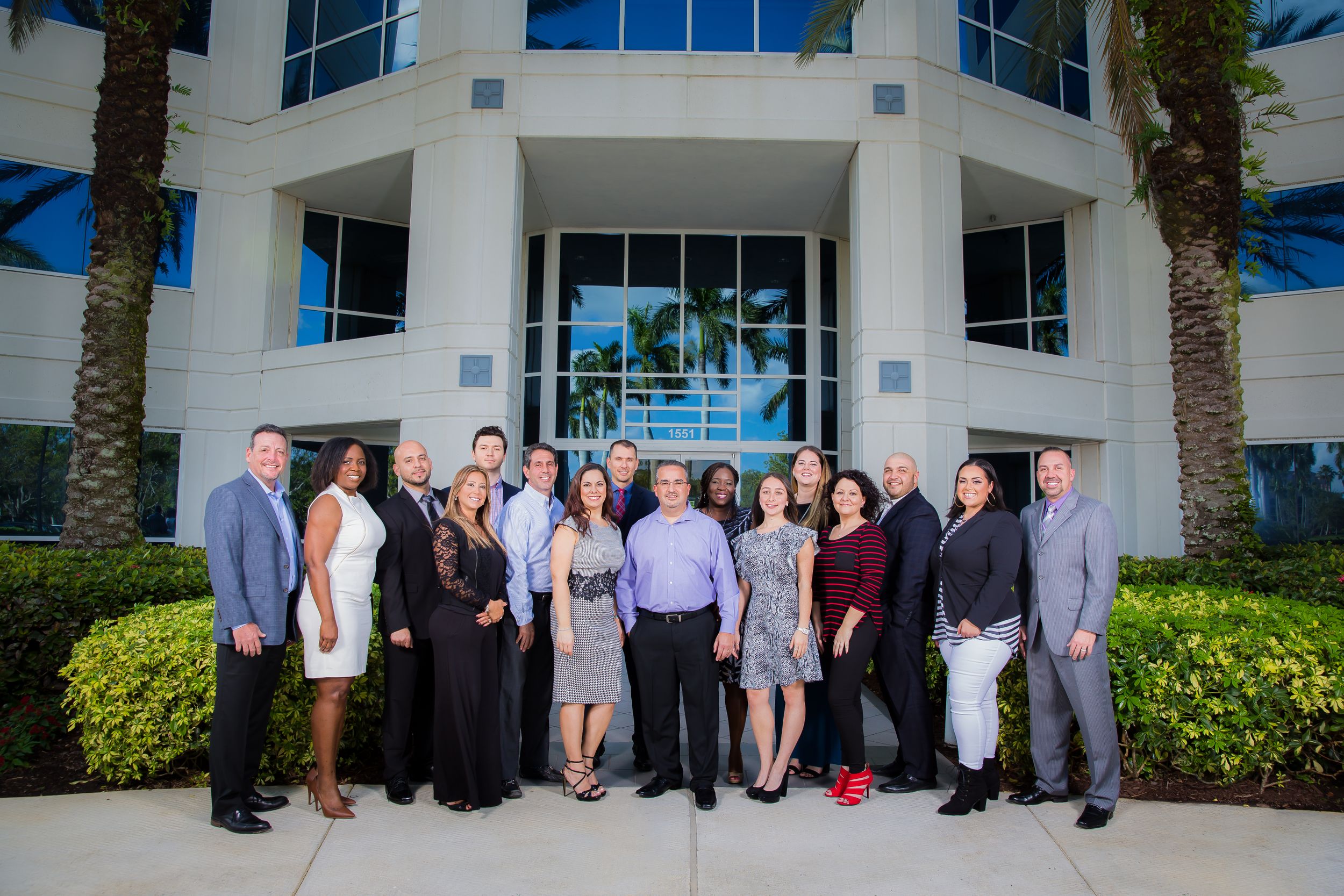 Professional business team members stand together outside a modern office building with palm trees.