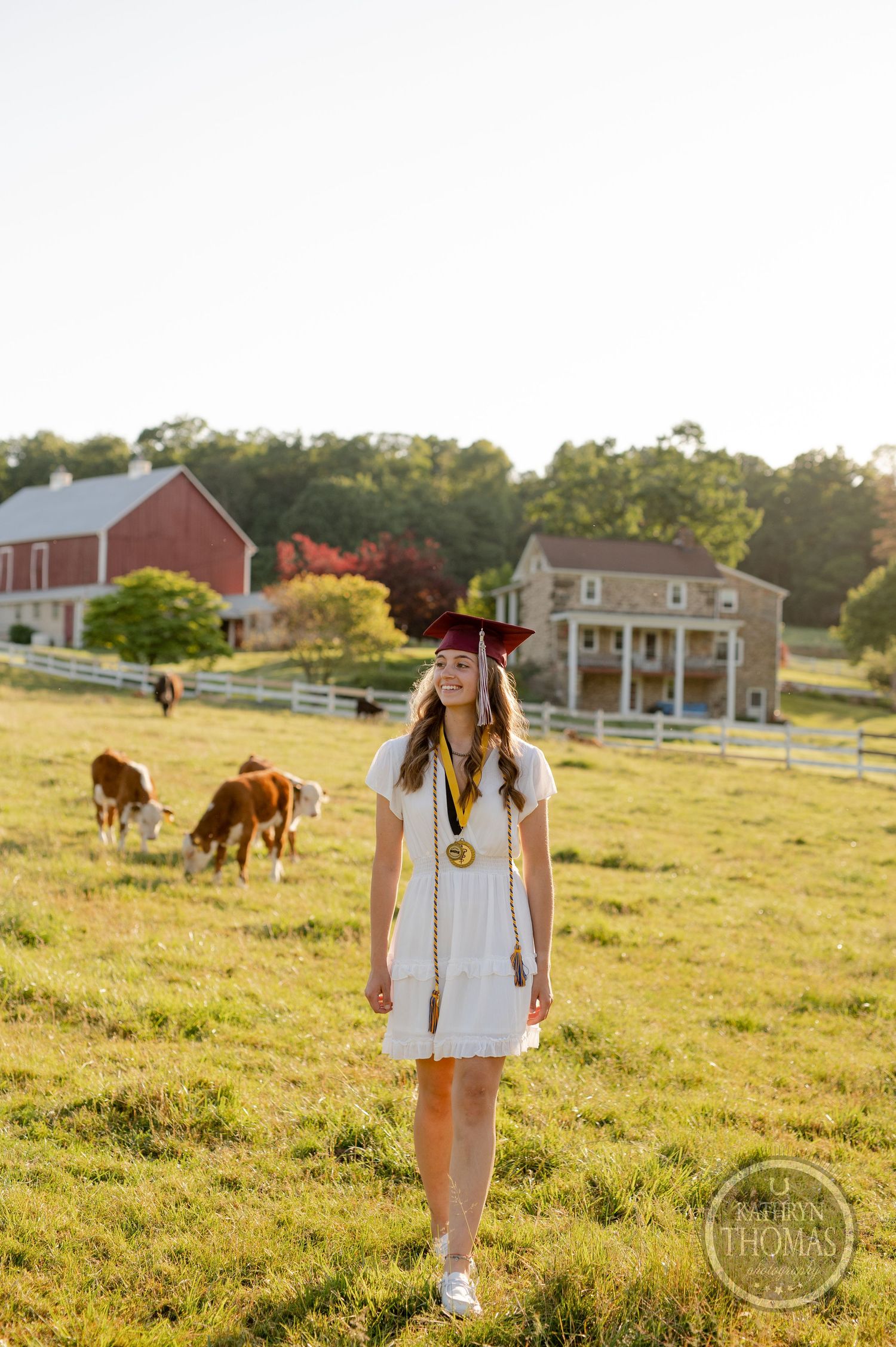 Graduate in white dress stands in sunny field with horses and red barn in background.