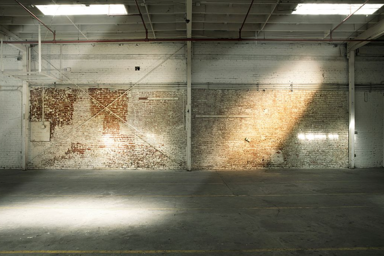 Sunlight streams across distressed warehouse wall creating dramatic shadows on concrete floor.