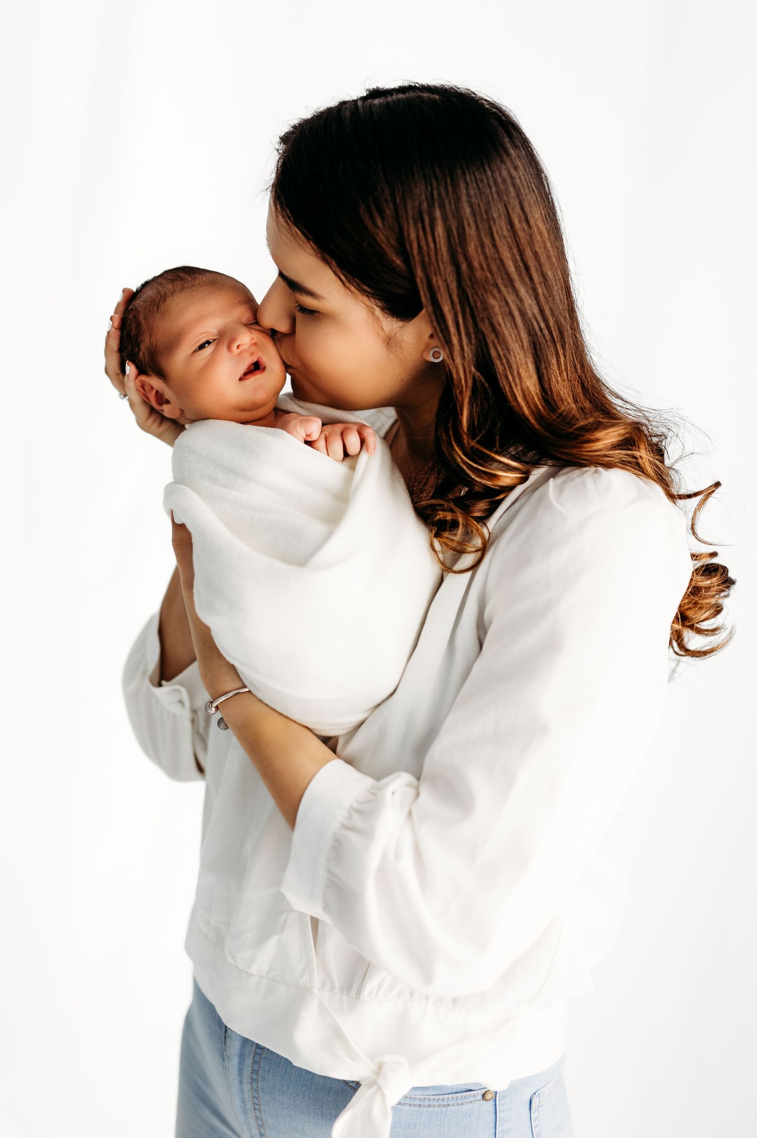 A tender moment as a mother in a white sweater kisses her swaddled newborn baby against a light background.
Rashinphotography-Nova DMV DC