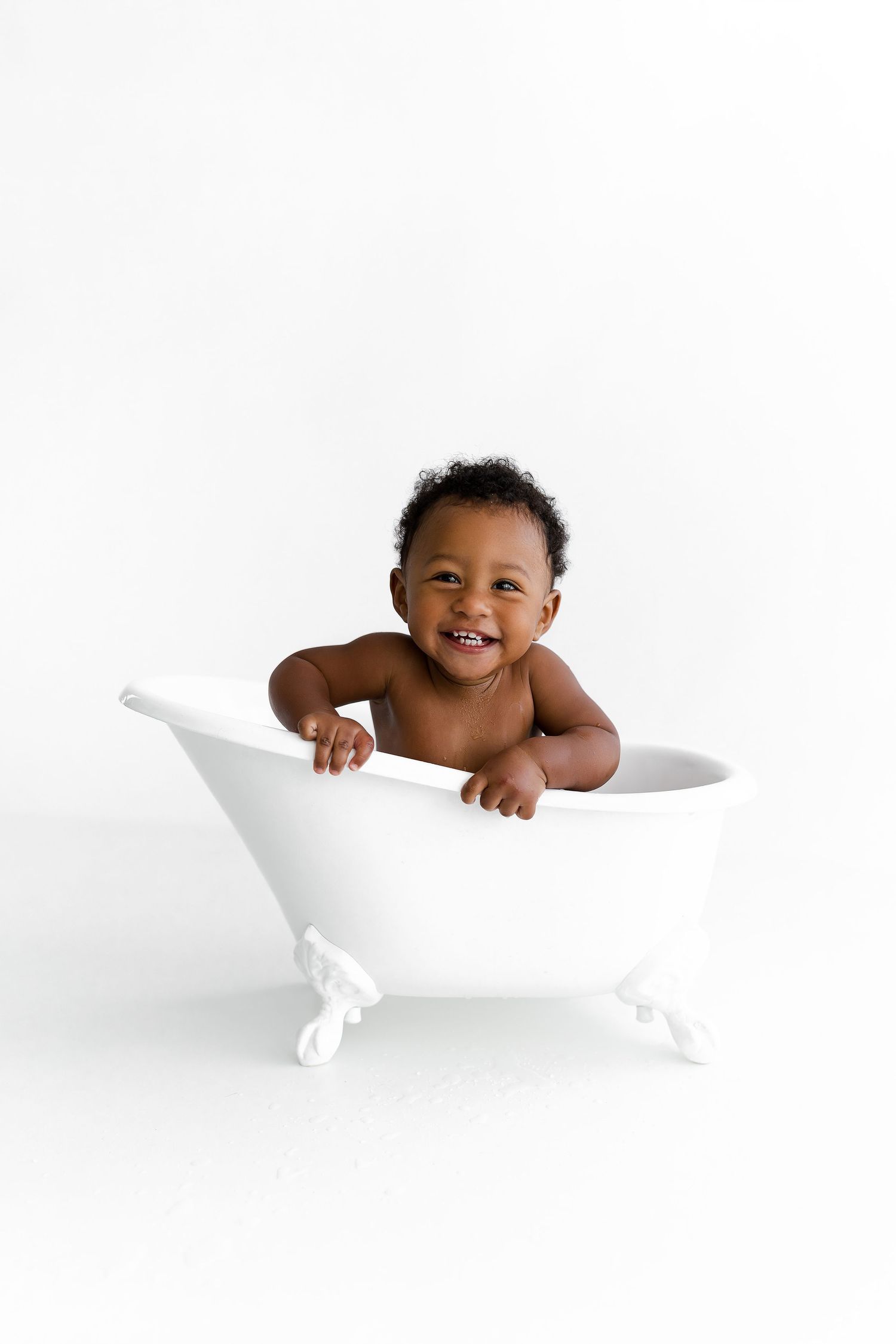 Happy baby sitting in white vintage clawfoot bathtub against white background during bath time photography session.